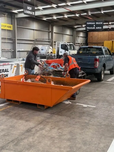 A tradie unloading into a skip in the tipping truck and trailer area, with a staff member helping